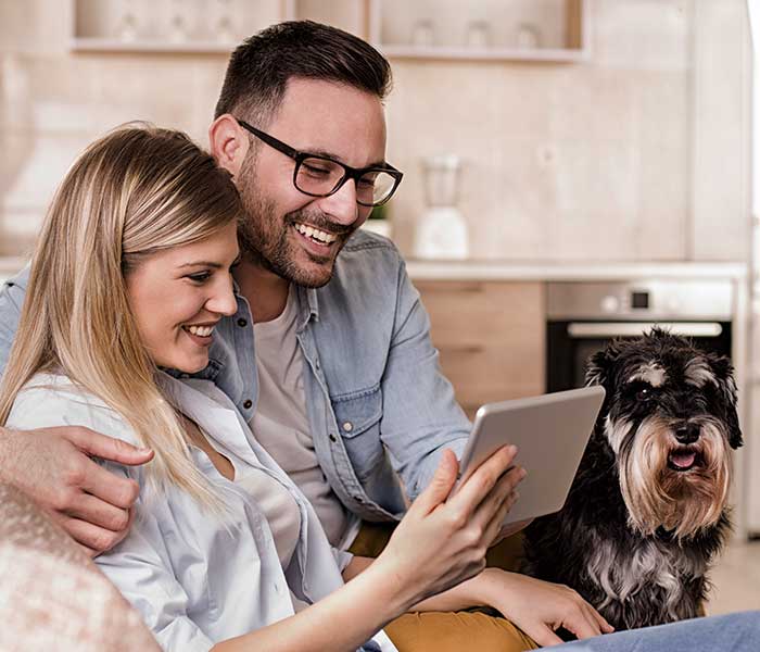 Young happy couple sitting on sofa in hug with dog and looking at tablet